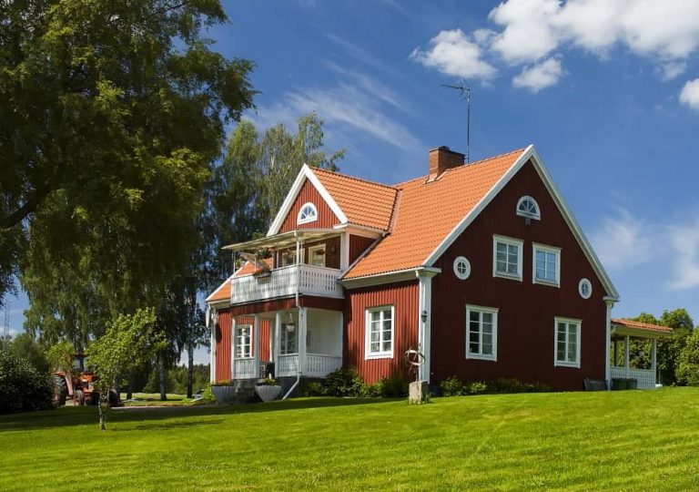 Rotes Schwedenhaus mit weißem Balkon, Satteldach und Holzfassade, umgeben von Rasen, Bäumen und ländlicher Natur unter blauem Himmel.