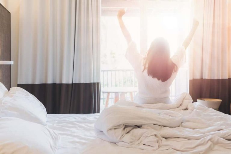 Frau sitzt im Bett und streckt sich morgens vor einem sonnendurchfluteten Fenster – Symbol für einen energiegeladenen Start in den Tag.