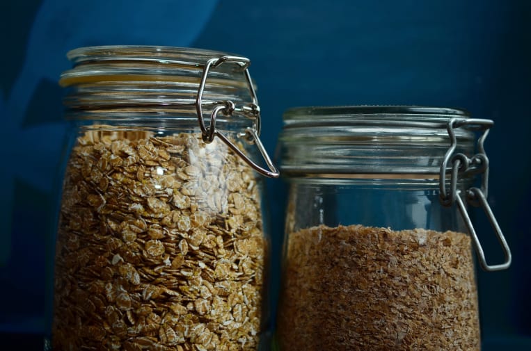 Zwei Einmachgläser mit Bügelverschluss auf einem Tisch. Das linke Glas ist mit Haferflocken gefüllt, während das rechte Glas gemahlenes Getreide enthält. Beide Gläser sind vor einem dunkelblauen Hintergrund platziert.