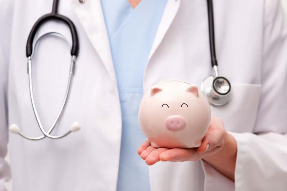 Ein Arzt in weißem Kittel und mit Stethoskop hält ein Sparschwein in der Hand. Das Sparschwein hat ein lächelndes Gesicht. Dies symbolisiert Verbindung zwischen Gesundheit und Finanzen.
