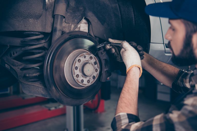 Ein Kfz-Mechaniker mit Handschuhen überprüft die Bremsscheibe eines Fahrzeugs mit einem Werkzeug, während das Auto auf einer Hebebühne steht. Im Bild sind die Aufhängung, die Federung und der Bremssattel gut sichtbar.