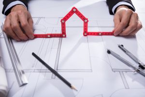 Architekt hält einen roten Gliedermaßstab in Form eines Hauses über Bauzeichnungen, umgeben von Lineal, Bleistift, Zirkel und Bauplänen auf einem Tisch.
