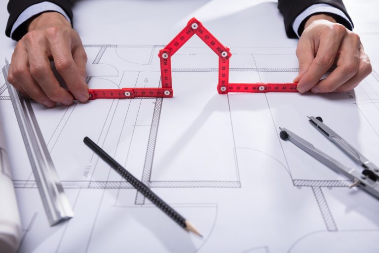 Architekt hält einen roten Gliedermaßstab in Form eines Hauses über Bauzeichnungen, umgeben von Lineal, Bleistift, Zirkel und Bauplänen auf einem Tisch.