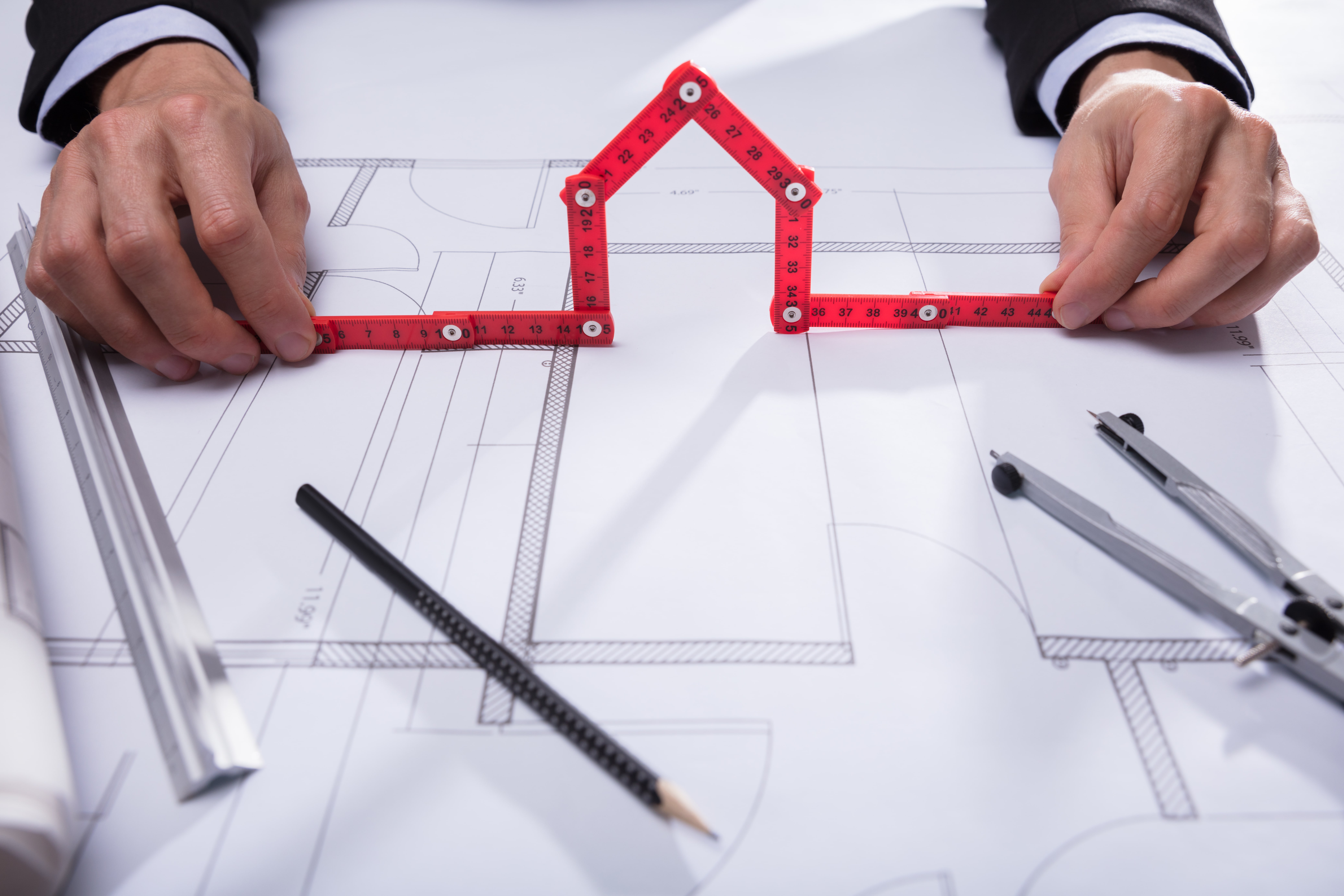 Architekt hält einen roten Gliedermaßstab in Form eines Hauses über Bauzeichnungen, umgeben von Lineal, Bleistift, Zirkel und Bauplänen auf einem Tisch.