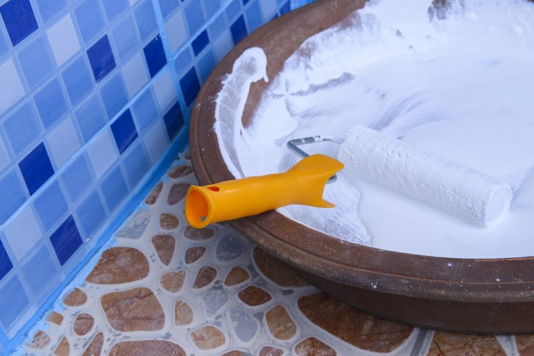Vorbereitung zum Streichen von Bodenfliesen im Badezimmer.. Farbwanne mit weißer Farbe und Farbrolle auf gemustertem Fliesenboden vor blauen Wandfliesen.