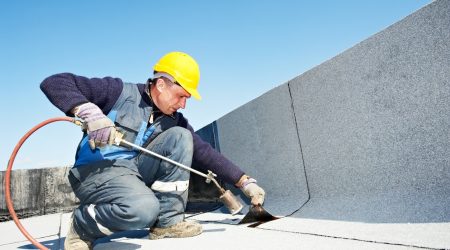 Dachdecker mit gelben Bauhelm verlegt Bitumenbahnen auf einem Dach