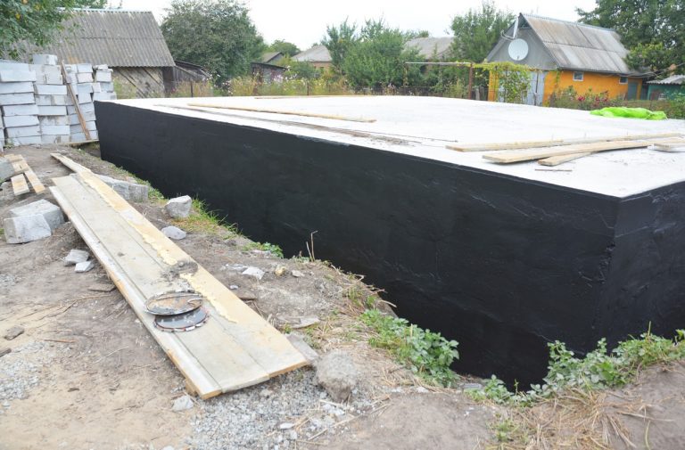 Frisch betoniertes und mit schwarzer Bitumenmasse abgedichtetes Hausfundament auf einer Baustelle, umgeben von Baumaterialien und Werkzeugen.