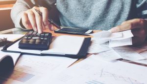 Person sitzt an einem Tisch mit Taschenrechner, Smartphone, Belegen und Dokumenten. Sie rechnet und notiert Zahlen im Rahmen einer Schenkungssteuer.