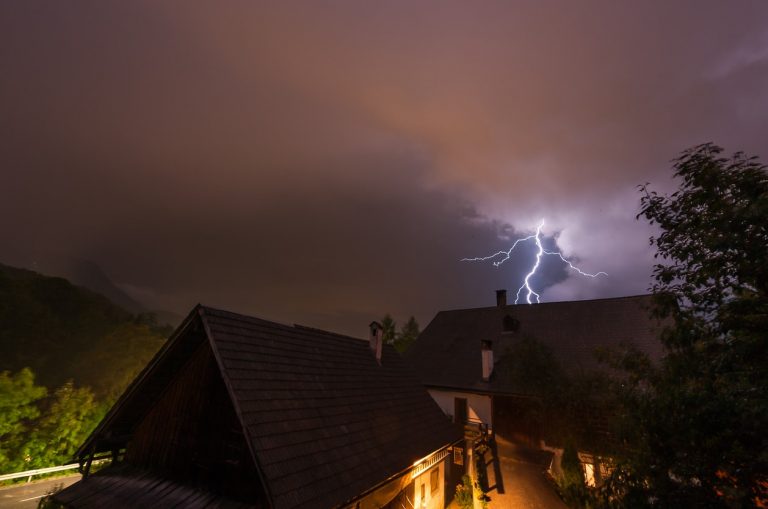 Nächtlicher Blitzeinschlag über Wohnhäusern in einer ländlichen Gegend; dramatischer Himmel mit dunklen Gewitterwolken und sichtbarer Entladung.