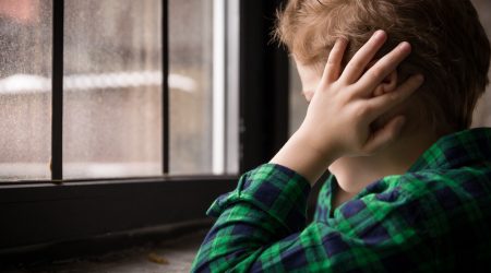 Kind sitzt mit den Händen an den Ohren vor einem Fenster mit einfachen Glasscheiben – Symbolbild für mangelhaften Schallschutz und Lärmbelastung in Wohnräumen.