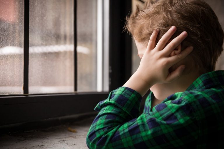 Kind sitzt mit den Händen an den Ohren vor einem Fenster mit einfachen Glasscheiben – Symbolbild für mangelhaften Schallschutz und Lärmbelastung in Wohnräumen.