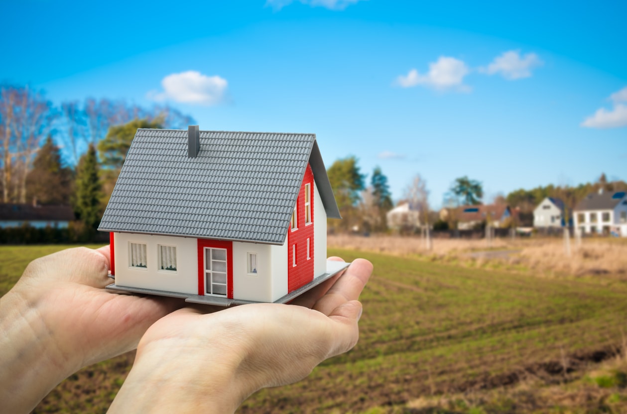 Kleines Haus in zwei Händen liegend vor ein potentielles Baugrundstück gehalten