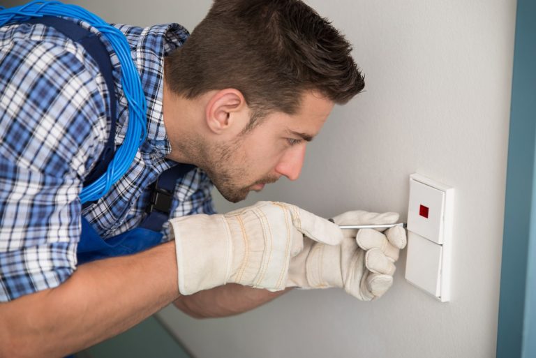 Ein Elektriker in kariertem Hemd und blauer Latzhose arbeitet an einem Wandschalter. Er trägt Arbeitshandschuhe und verwendet einen Schraubenzieher, während ein Kabel um seine Schulter hängt.
