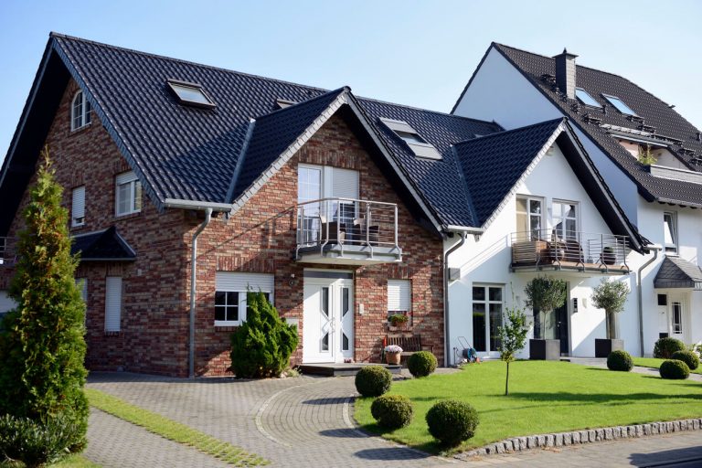 Modernes Mehrfamilienhaus mit Kombination aus Klinker- und Putzfassade, Balkonen, gepflegtem Vorgarten und dunklem Satteldach bei Sonnenschein.