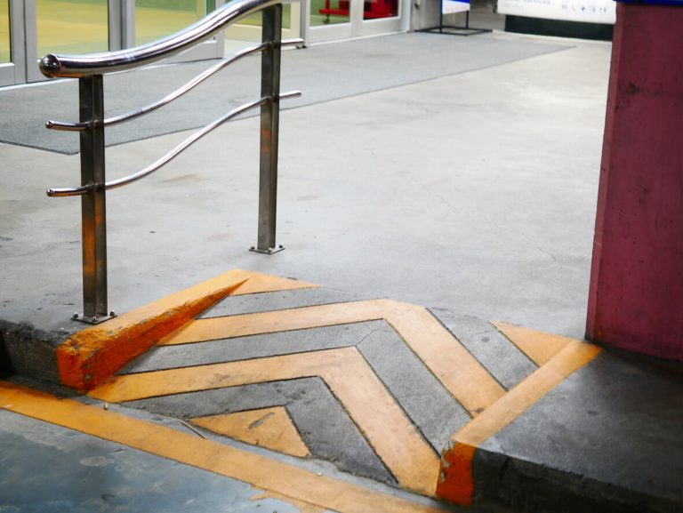 Barrierefreie Rampe mit gelb-schwarzen Markierungen und Edelstahl-Handlauf in einem öffentlichen Gebäude, z. B. in einem Einkaufszentrum oder Parkhaus.