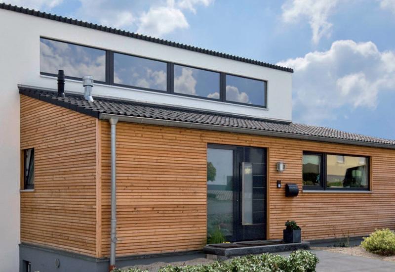 Moderner Hausanbau mit flacher Dachform, großflächigen Fenstern und Holzverschalung – Kombination aus natürlichen Materialien und klarer Linienführung. Ein Beispielhaus eines Holzrahmenbaus der Firma Puschmann Architektur.