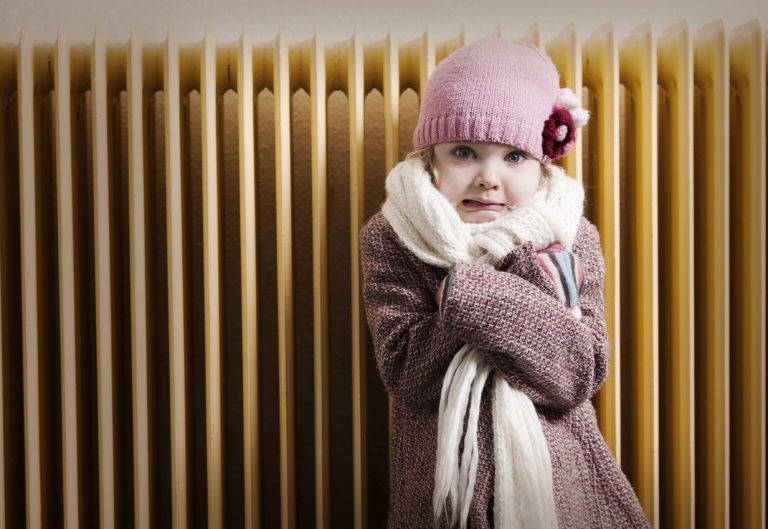 Kleines Mädchen friert mit Jacke, Mütze und Schal vor einer kalten Heizung