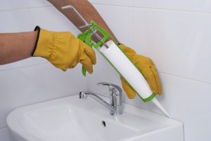 Ein Handwerker trägt gelbe Handschuhe und benutzt eine grüne Kartuschenpistole, um Silikon entlang der Kante eines weißen Waschbeckens auf weiße Fliesen aufzutragen, um so die Fugen zu erneuern.
