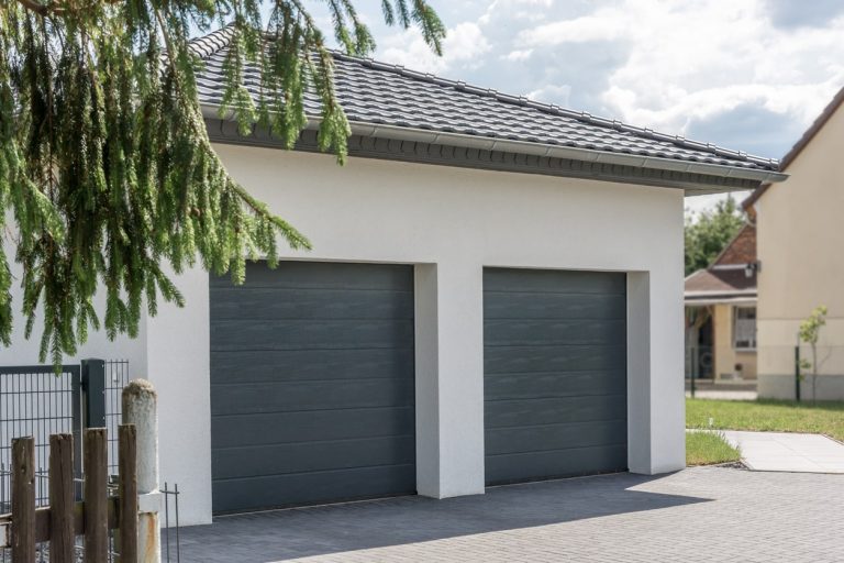 Moderne Fertig-Doppelgarage mit zwei anthrazitfarbenen Sektionaltoren, Flachdach mit Dachziegeln und gepflasterter Einfahrt, in einer Wohnsiedlung bei Tageslicht.