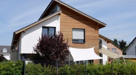 Modernes Wohnhaus mit asymmetrischem Pultdach, weißer Putzfassade und Holzverschalung, davor Sonnensegel und Gartenbepflanzung.