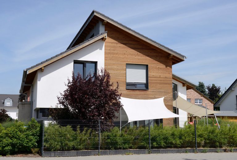 Modernes Wohnhaus mit asymmetrischem Pultdach, weißer Putzfassade und Holzverschalung, davor Sonnensegel und Gartenbepflanzung.