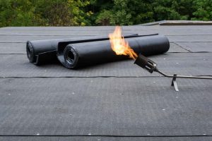 Zwei gerollte Bitumenbahnen und ein Propangasbrenner mit offener Flamme auf einem Flachdach, bereit zur Dachabdichtung durch Verschweißen.