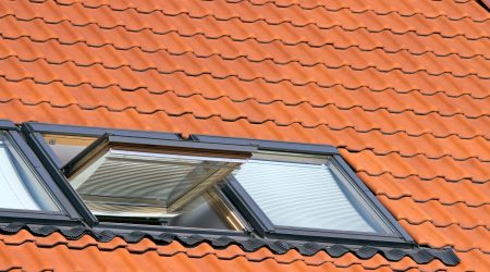 Zwei moderne Dachfenster mit integriertem Sonnenschutz in ein rotes Ziegeldach eingelassen, eines davon gekippt geöffnet, das andere geschlossen.