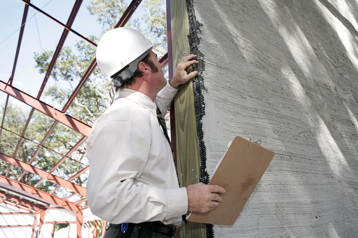 Baugutachter mit weißem Schutzhelm prüft Wand mit Checkliste bei einer Bauabnahme