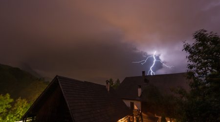 Nächtliche Szene in einem Dorf mit dunklen Dächern, im Hintergrund zuckender Blitz am Himmel über bewaldeten Hügeln. Die Gewitterwolken dominieren den Himmel, während der Blitz das Umfeld dramatisch erhellt.
