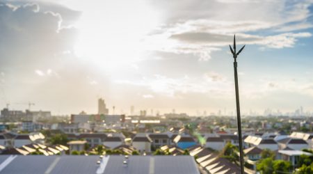 Blick über eine Siedlung mit einem Blitzableiter im Vordergrund auf einem Flachdach. Der Blitzschutz besteht aus einer vertikal montierten Metallstange mit mehrfacher Spitze. Im Hintergrund erstreckt sich eine dichte Stadtlandschaft bei Sonnenuntergang.