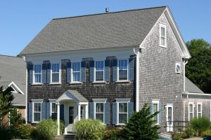 Zweigeschossiges Einfamilienhaus mit dunkelgrauen Holzschindeln an der Fassade und einem Satteldach mit Dachschindeln. Klassisches amerikanisches Design mit weißen Fensterrahmen und blauen Fensterläden.