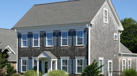 Zweigeschossiges Einfamilienhaus mit dunkelgrauen Holzschindeln an der Fassade und einem Satteldach mit Dachschindeln. Klassisches amerikanisches Design mit weißen Fensterrahmen und blauen Fensterläden.