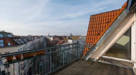 Städtische Dachterrasse mit Holzbelag und Blumenkästen am Geländer, Blick auf eine lange Häuserzeile mit roten Ziegeldächern und einer urbanen Skyline im Hintergrund.