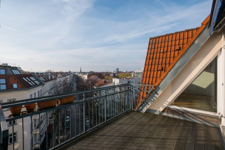Städtische Dachterrasse mit Holzbelag und Blumenkästen am Geländer, Blick auf eine lange Häuserzeile mit roten Ziegeldächern und einer urbanen Skyline im Hintergrund.