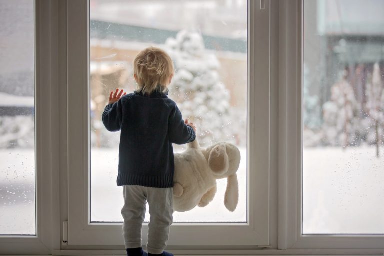 Kleines Kind steht mit Kuscheltier im Arm vor Fenster aus Isolierglas und schaut in den verschneiten Garten