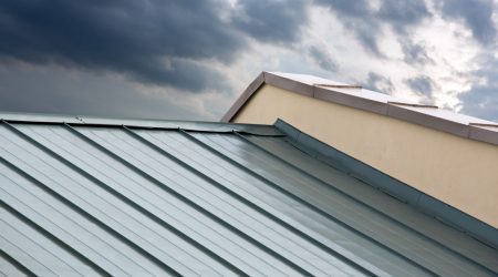 Modernes Dach mit Stehfalzdeckung aus Metall bei bewölktem Himmel. Die klaren Linien der Blechbahnen verlaufen diagonal über das geneigte Dach eines hell verputzten Gebäudes.