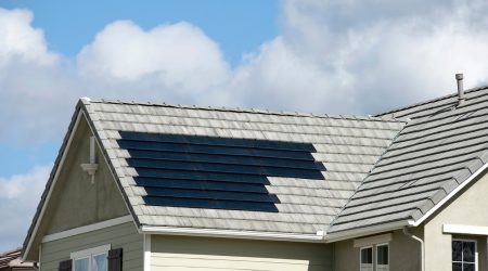 Einfamilienhaus mit hellgrauem Ziegeldach, in das flächenbündig schwarze Solarziegel integriert sind, bei leicht bewölktem Himmel.