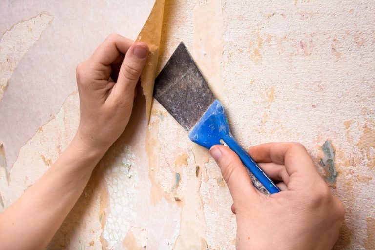 Frauenhände kratzen mit blauer Spachtel alte Tapete von einer Wand