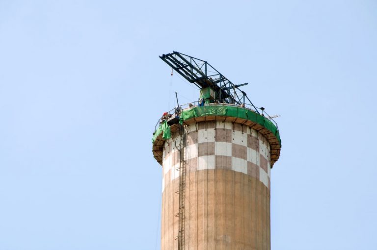 Konsolgerüst auf einem hohen Industrieschornstein mit Kranaufbau und Sicherungsnetz für Sanierungsarbeiten unter freiem Himmel.