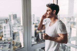 Junger Mann mit Kaffeetasse telefoniert am Fenster einer modernen Wohnung mit Blick auf die Stadt – Symbolbild für Lärmschutz durch Schallschutzglas.
