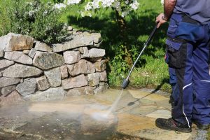 Mann mit blauer Arbeitshose reinigt mit Hochdruckreiniger Steinplatten einer Terrasse