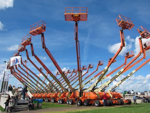 Mehrere orange Teleskoparbeitsbühnen in aufgerichteter Position auf einer Messefläche unter blauem Himmel.