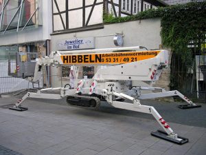 Kompakte Raupenarbeitsbühne mit ausgefahrenen Stützfüßen vor einem Juweliergeschäft, Arbeitsbühnenvermietung Hibbeln mit Telefonnummer auf dem Ausleger.