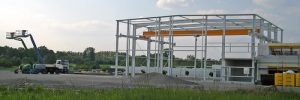 Stahlkonstruktion einer Industriehalle im Rohbau auf einer Baustelle im Grünen, mit Hebebühne und Baumaterialien im Vordergrund.