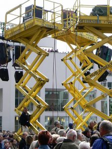 Zwei gelbe Scherenarbeitsbühnen bei einer Veranstaltung im Freien, umgeben von einer Menschenmenge, mit montierter Licht- und Tontechnik.