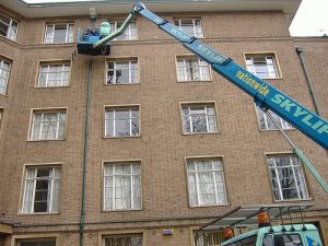Arbeiter auf einer blauen Hebebühne bei Wartungsarbeiten an der Fassade eines mehrstöckigen Backsteingebäudes.