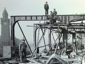 Schwarzweißaufnahme von Arbeitern auf einer Eisenbrücke während des B Conruhs, im Hintergrund ein Uhrturm und Baugerüste aus Stahlträgern.