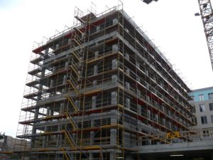 Großflächiges Fassadengerüst an einem mehrstöckigen Rohbau mit integrierter Gerüsttreppe aus gelben Stahlbauteilen zur sicheren vertikalen Erschließung während der Bauarbeiten.