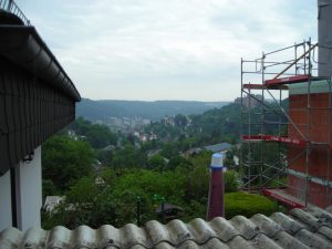 Teil eines Fassadengerüsts an einem Gebäude mit Blick über eine grüne Hügellandschaft und Stadt im Tal bei bewölktem Himmel.