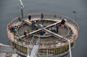 Bauarbeiter auf einer runden Schalung über Wasser beim Bewehren und Betonieren eines Brückenfundaments mit Arbeitsschutz und Zugang über Gerüsttreppe.